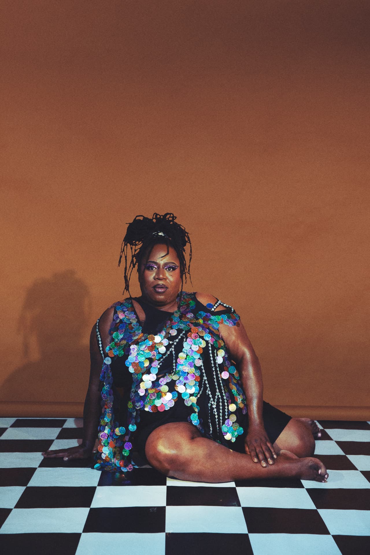Terrie Talks seated on a checkerboard floor in a sequinned outfit against a warm brown backdrop